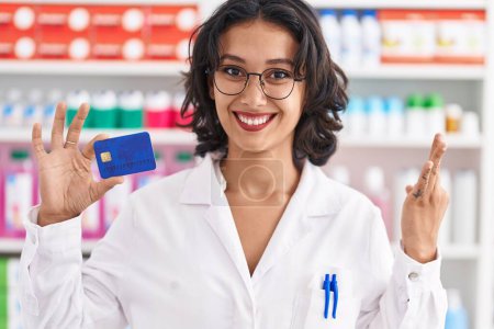 Photo for Young hispanic woman working at pharmacy drugstore holding credit card doing ok sign with fingers, smiling friendly gesturing excellent symbol - Royalty Free Image