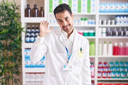 Foto de Hombre hispano guapo trabajando en farmacia renunciando a decir hola feliz y sonriente, gesto de bienvenida amistoso - Imagen libre de derechos