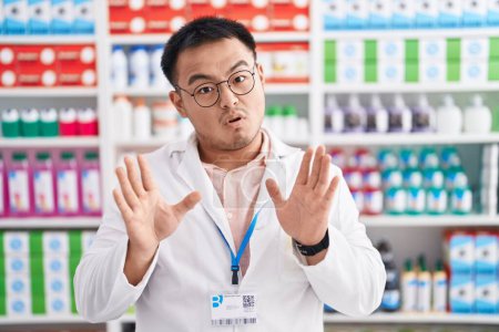 Photo for Chinese young man working at pharmacy drugstore moving away hands palms showing refusal and denial with afraid and disgusting expression. stop and forbidden. - Royalty Free Image