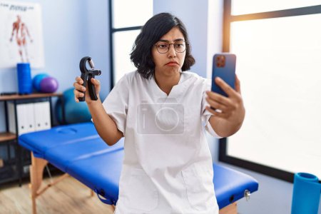 Foto de Young hispanic physiotherapist woman holding hand grip to train muscle doing video call depressed and worry for distress, crying angry and afraid. sad expression. - Imagen libre de derechos