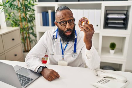 Foto de Hombre afroamericano trabajando en clínica dietista sosteniendo donut asustado y sorprendido con la boca abierta para sorpresa, cara de incredulidad - Imagen libre de derechos
