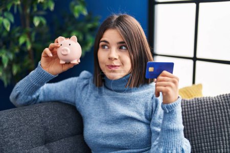 Foto de Mujer hispana joven sosteniendo alcancía y tarjeta de crédito sonriendo mirando hacia un lado y mirando hacia otro pensando. - Imagen libre de derechos