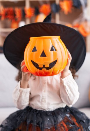 Photo for Adorable hispanic girl wearing halloween costume holding pumpkin basket over face at home - Royalty Free Image