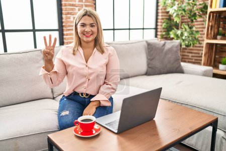 Foto de Mujer hispana joven usando portátil sentado en el sofá en casa mostrando y apuntando hacia arriba con los dedos número tres mientras sonríe confiado y feliz. - Imagen libre de derechos