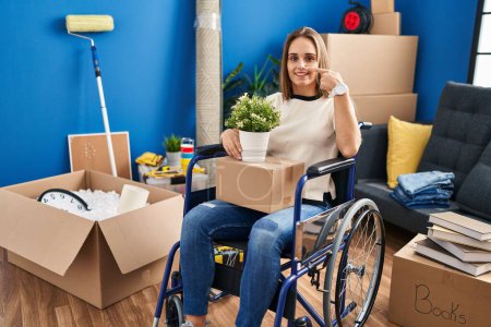 Téléchargez les photos : Jeune femme assise en fauteuil roulant se déplaçant vers une nouvelle maison pointant du doigt et du nez, souriante et joyeuse. concept de beauté - en image libre de droit