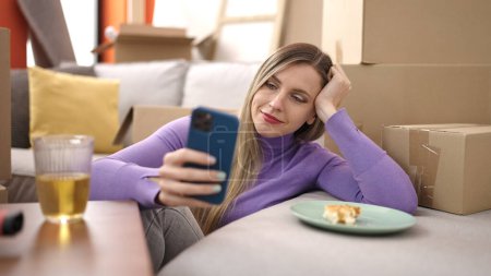 Téléchargez les photos : Jeune femme blonde utilisant smartphone assis sur le sol à la nouvelle maison - en image libre de droit