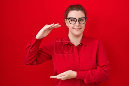 Photo for Young hispanic woman with red hair standing over red background gesturing with hands showing big and large size sign, measure symbol. smiling looking at the camera. measuring concept. - Royalty Free Image