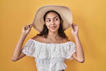 Foto de Mujer brasileña joven con sombrero de verano sobre fondo amarillo sonriendo mirando hacia un lado y mirando hacia otro pensando. - Imagen libre de derechos