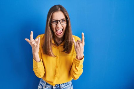 Téléchargez les photos : Jeune femme portant des lunettes debout sur fond bleu criant avec une expression folle faire symbole rock avec les mains en l'air. star de la musique. concept lourd. - en image libre de droit