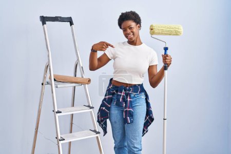 Foto de Mujer afroamericana sosteniendo rodillo pintor mirando confiado con sonrisa en la cara, señalándose con los dedos orgullosos y felices. - Imagen libre de derechos