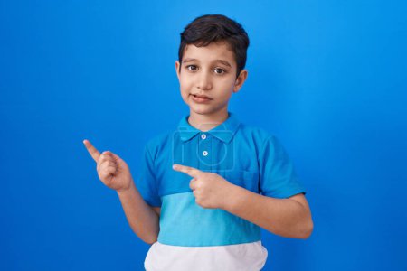 Foto de Pequeño niño hispano de pie sobre fondo azul sonriendo y mirando a la cámara apuntando con dos manos y dedos hacia un lado. - Imagen libre de derechos