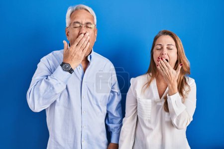 Photo for Middle age hispanic couple standing over blue background bored yawning tired covering mouth with hand. restless and sleepiness. - Royalty Free Image