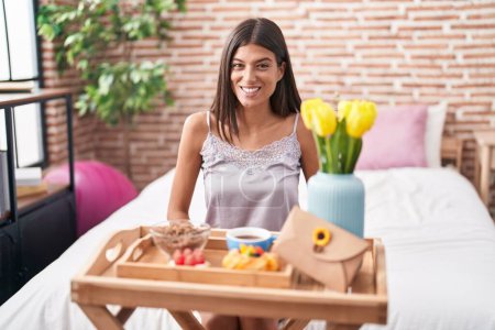 Téléchargez les photos : Brunette young woman eating breakfast sitting on the bed looking positive and happy standing and smiling with a confident smile showing teeth - en image libre de droit