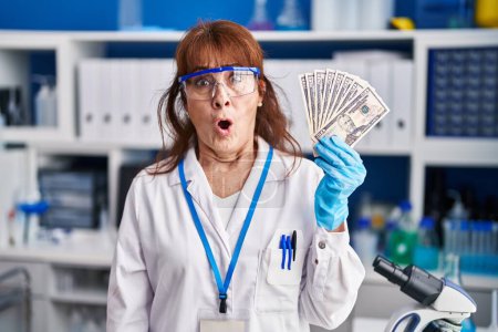Foto de Middle age hispanic woman working at scientist laboratory holding dollars scared and amazed with open mouth for surprise, disbelief face - Imagen libre de derechos