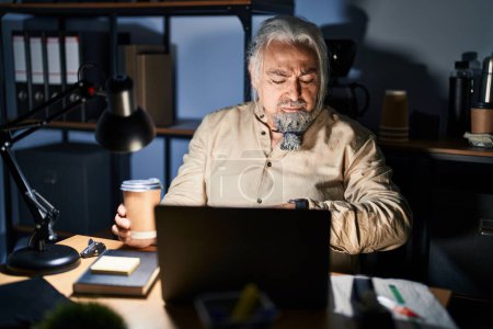 Téléchargez les photos : Homme d'âge moyen aux cheveux gris travaillant au bureau la nuit vérifiant l'heure sur la montre-bracelet, détendu et confiant - en image libre de droit