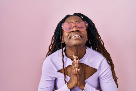 Téléchargez les photos : Femme africaine avec les cheveux tressés debout sur fond rose mendier et prier avec les mains ainsi que l'expression de l'espoir sur le visage très émotionnel et inquiet. mendier. - en image libre de droit