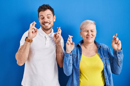 Téléchargez les photos : Young brazilian mother and son standing over blue background gesturing finger crossed smiling with hope and eyes closed. luck and superstitious concept. - en image libre de droit