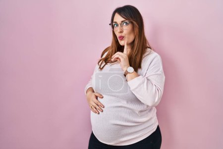 Foto de Pregnant woman standing over pink background thinking concentrated about doubt with finger on chin and looking up wondering - Imagen libre de derechos