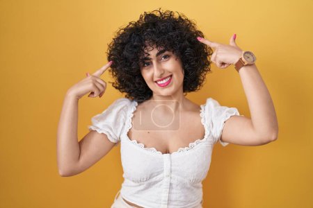 Photo for Young brunette woman with curly hair standing over yellow background smiling pointing to head with both hands finger, great idea or thought, good memory - Royalty Free Image