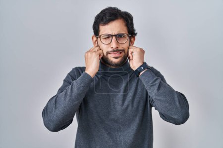 Foto de Hombre latino guapo de pie sobre un fondo aislado cubriendo las orejas con dedos con expresión molesta por el ruido de la música fuerte. concepto de sordo. - Imagen libre de derechos