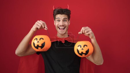 Photo for Young hispanic man wearing devil costume holding halloween pumpkin baskets over isolated red background - Royalty Free Image