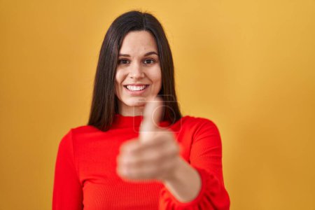 Foto de Mujer hispana joven de pie sobre fondo amarillo sonriente amistoso ofreciendo apretón de manos como saludo y bienvenida. negocios exitosos. - Imagen libre de derechos