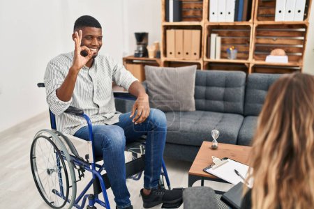 Foto de Hombre afroamericano haciendo terapia sentado en silla de ruedas sonriendo positivo haciendo signo de bien con la mano y los dedos. expresión exitosa. - Imagen libre de derechos