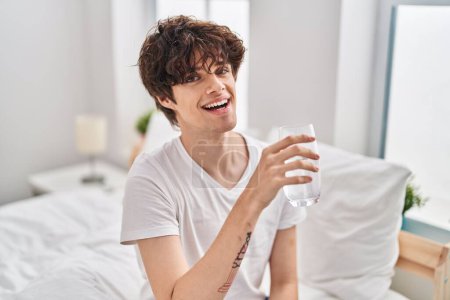 Foto de Joven hispano bebiendo vaso de agua sentado en la cama en el dormitorio - Imagen libre de derechos