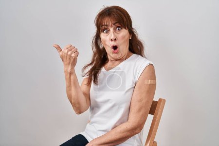 Photo for Middle age hispanic woman getting vaccine showing arm with band aid surprised pointing with hand finger to the side, open mouth amazed expression. - Royalty Free Image