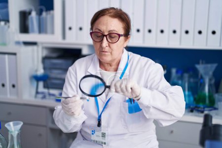 Foto de Senior woman scientist using magnifying glass holding pill at laboratory - Imagen libre de derechos