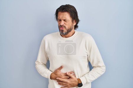 Foto de Handsome middle age man wearing casual sweater over blue background with hand on stomach because nausea, painful disease feeling unwell. ache concept. - Imagen libre de derechos