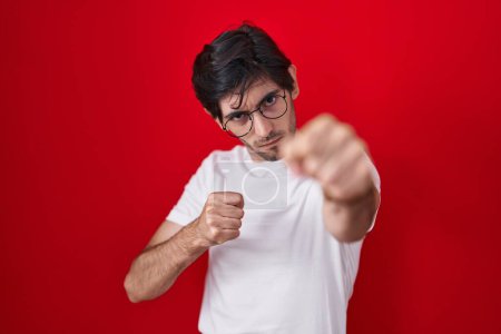 Photo for Young hispanic man standing over red background punching fist to fight, aggressive and angry attack, threat and violence - Royalty Free Image