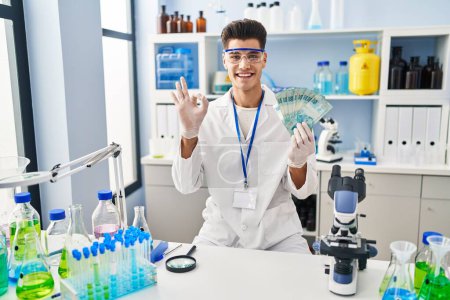 Foto de Joven hombre hispano trabajando en laboratorio científico sosteniendo reales brasileños haciendo signo de ok con los dedos, sonriendo gesto amistoso excelente símbolo - Imagen libre de derechos