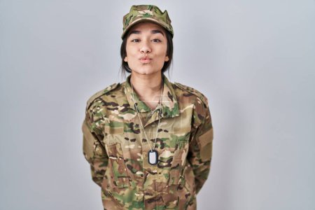 Foto de Young south asian woman wearing camouflage army uniform looking at the camera blowing a kiss on air being lovely and sexy. love expression. - Imagen libre de derechos