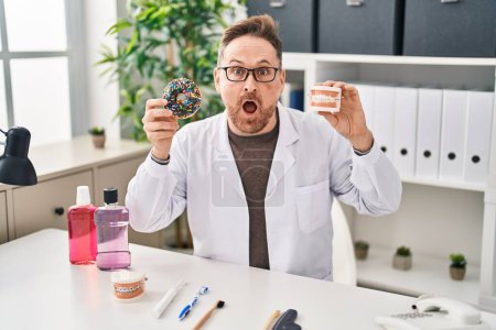 Photo for Middle age caucasian dentist man holding denture and doughnuts afraid and shocked with surprise and amazed expression, fear and excited face. - Royalty Free Image