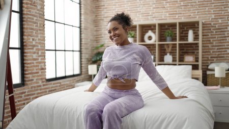 Téléchargez les photos : Femme afro-américaine souriante confiante assise sur le lit dans la chambre - en image libre de droit
