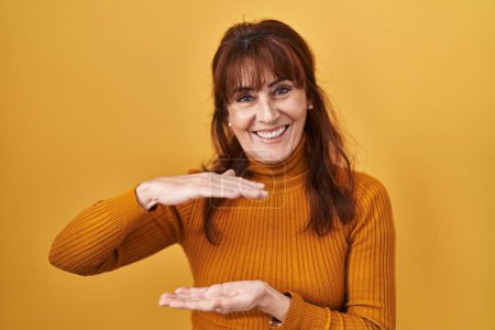 Téléchargez les photos : Middle age hispanic woman standing over yellow background gesturing with hands showing big and large size sign, measure symbol. smiling looking at the camera. measuring concept. - en image libre de droit