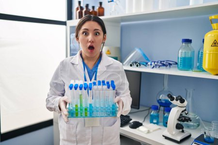 Téléchargez les photos : Young latin woman working at scientist laboratory afraid and shocked with surprise and amazed expression, fear and excited face. - en image libre de droit