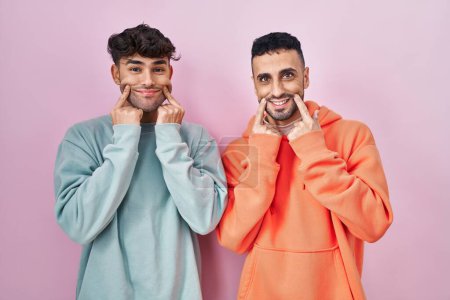 Foto de Joven pareja gay hispana de pie sobre fondo rosa sonriendo con la boca abierta, los dedos señalando y forzando sonrisa alegre - Imagen libre de derechos