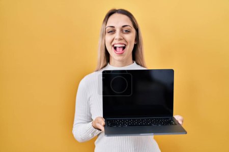 Foto de Joven mujer rubia sosteniendo portátil sonriendo y riendo en voz alta porque broma loca divertida. - Imagen libre de derechos