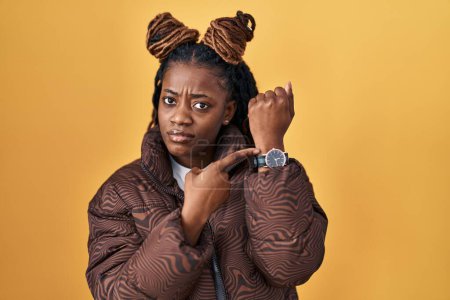 Téléchargez les photos : Femme africaine avec les cheveux tressés debout sur fond jaune à la hâte pointant vers regarder le temps, l'impatience, regardant la caméra avec une expression détendue - en image libre de droit
