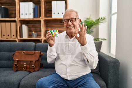 Foto de Senior therapist with grey hair playing colorful puzzle cube intelligence game doing ok sign with fingers, smiling friendly gesturing excellent symbol - Imagen libre de derechos