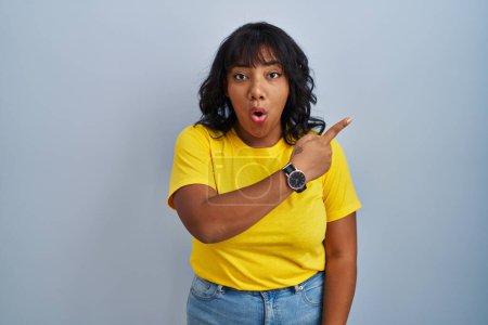 Photo for Hispanic woman standing over blue background surprised pointing with finger to the side, open mouth amazed expression. - Royalty Free Image