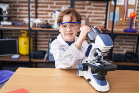 Téléchargez les photos : Adorable garçon hispanique étudiant souriant confiant en utilisant le microscope à la salle de classe de laboratoire - en image libre de droit