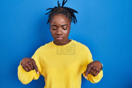 Téléchargez les photos : Belle femme noire debout sur fond bleu pointant vers le bas regardant triste et contrarié, indiquant la direction avec les doigts, malheureux et déprimé. - en image libre de droit