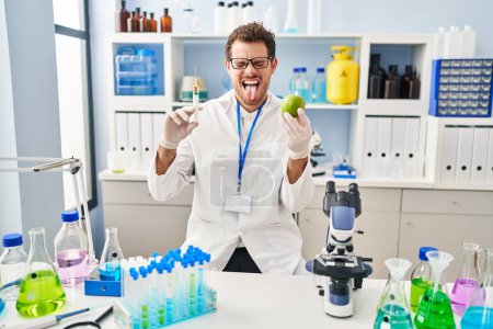 Foto de Young hispanic man working at scientist laboratory holding apple sticking tongue out happy with funny expression. - Imagen libre de derechos