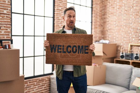 Foto de Middle age caucasian man holding welcome doormat at new home in shock face, looking skeptical and sarcastic, surprised with open mouth - Imagen libre de derechos