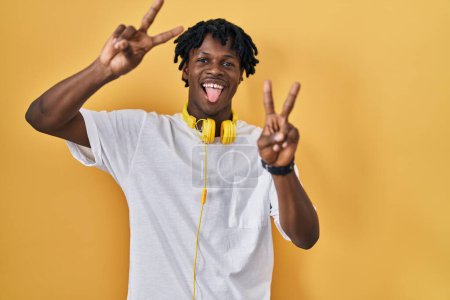 Photo for Young african man with dreadlocks standing over yellow background smiling with tongue out showing fingers of both hands doing victory sign. number two. - Royalty Free Image