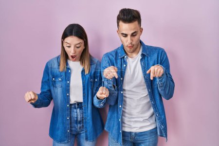 Téléchargez les photos : Jeune couple hispanique debout sur fond rose pointant vers le bas avec les doigts montrant la publicité, visage surpris et bouche ouverte - en image libre de droit