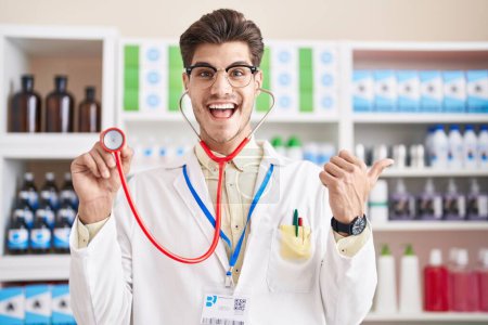 Téléchargez les photos : Young hispanic man working at pharmacy drugstore using stethoscope pointing thumb up to the side smiling happy with open mouth - en image libre de droit